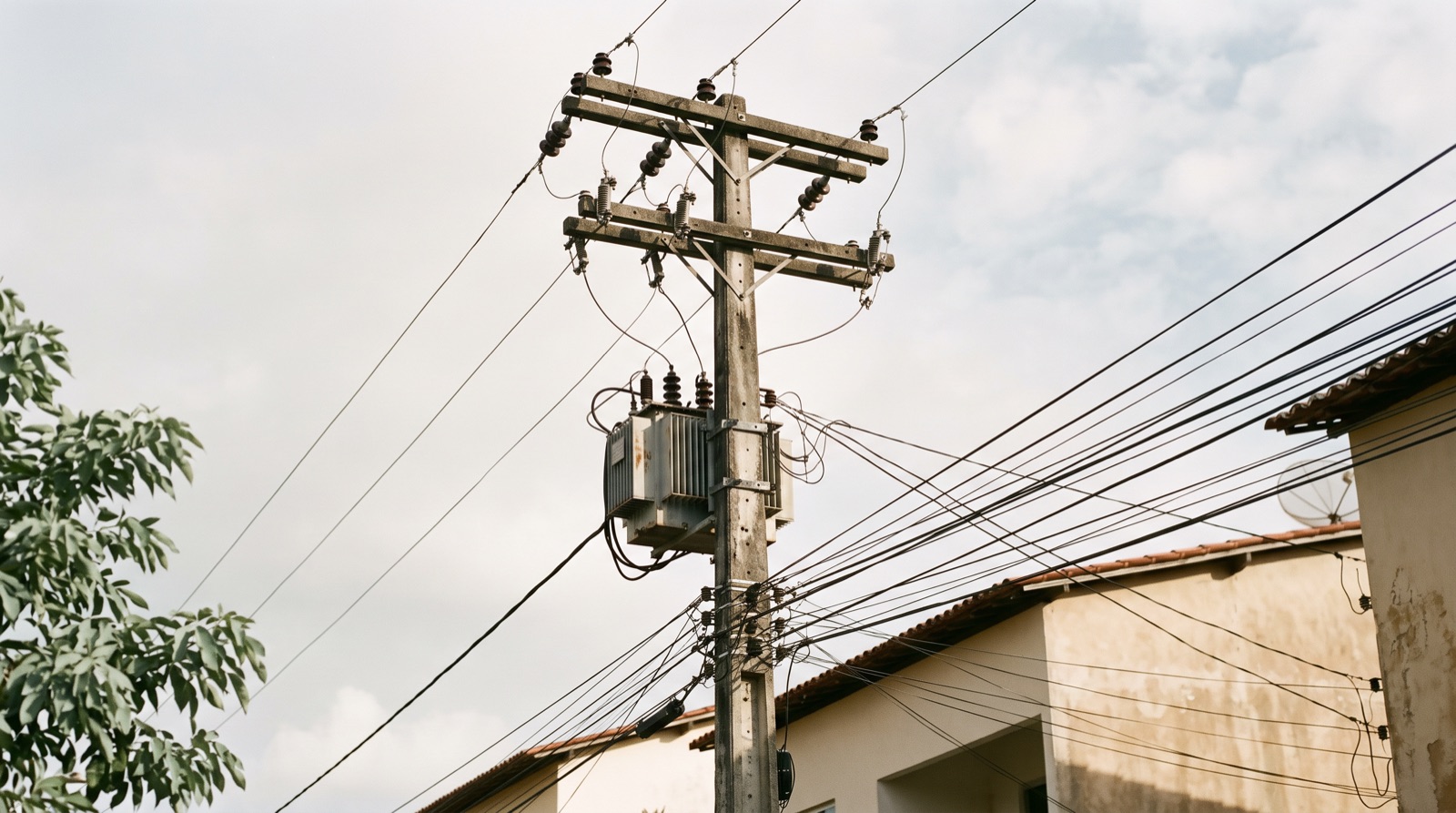 Poste de concreto com transformador e um emaranhado de fios de distribuição em rua residencial do Nordeste, com telhado de telha cerâmica ao lado.